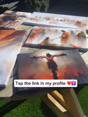 A table displays several paintings of religious scenes. The paintings are on canvas and depict figures in various settings, painted in a style that blends realism with artistic interpretation. One painting prominently displays a figure on a cross, set against a burning background, which hints at somberness. Another picture involves figures in orange garments. The paintings are atop a wooden table placed outside. A text overlay, placed at the bottom, features white text in a rounded rectangle with a heart and a cross symbol.