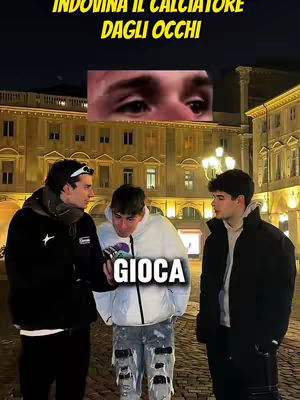 The image shows three young men standing on a brick-paved area in front of a building at night. The text overlay reads, "INDOVINA IL CALCIATORE DAGLI OCCHI" (Guess the footballer from the eyes) above a cropped image of a person's eyes. Below this, a text box at the bottom with the word "GIOCA" (Play). The men wear dark jackets and are positioned to fill the frame, suggesting a game setup.