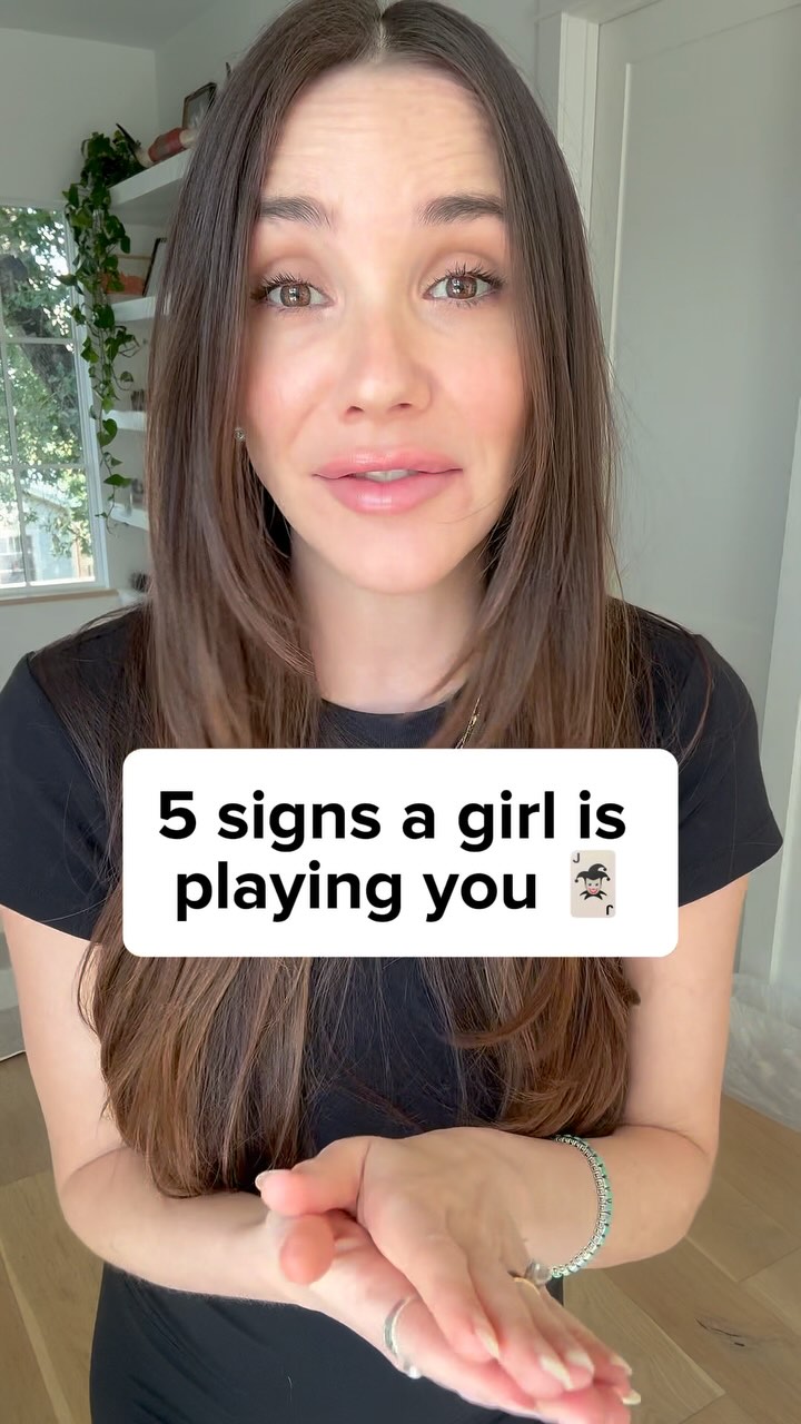 A woman with long brown hair is centered in the frame, looking directly at the camera. She has olive skin, with a white text overlay on her face. The background is a softly lit room with neutral tones and a hint of greenery in the corner.