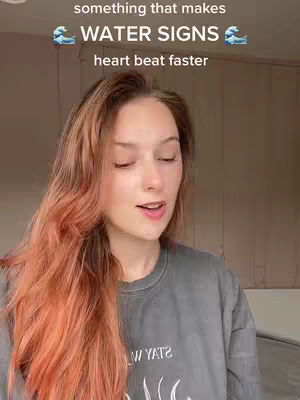 A woman with flowing auburn hair is centered in the frame. She is wearing a gray shirt. Above her head is text overlay in a white font with a wave emoji. The background is a muted beige wall.