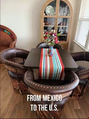 The image features a dining setup with a table and chairs. A dark brown square table is set with a colorful striped table runner and a vase of flowers. Surrounding the table are chairs made of dark brown leather and woven wood. In the background, there is a wooden cabinet with glass doors, a small window, and a portion of a leather chair. The overall composition has warm tones and is well-lit, suggesting a cozy interior.