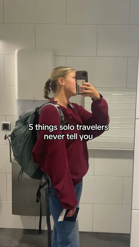 A woman in a burgundy sweater and jeans stands in a bathroom, holding a phone up to take a selfie. She has a backpack on. Text overlay says "5 things solo travelers never tell you."