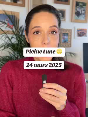 A woman with brown hair and eyes is centered in the frame. She is wearing a red sweater and appears to be holding a small, black microphone in her right hand. Above her head are two text boxes, and behind her there is a background of framed pictures and a green plant.