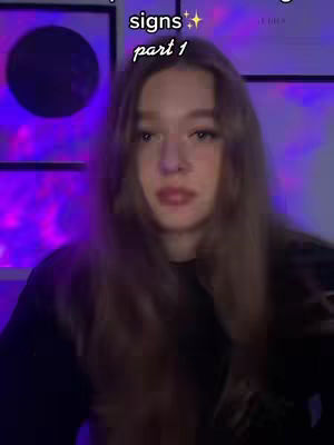 A young woman with long brown hair is centered in the frame. She is wearing a black top. The background is dimly lit, with purple and blue hues. Text overlayed on the image says 'rising signs' and 'part 1'. The overall composition suggests a casual, personal video.