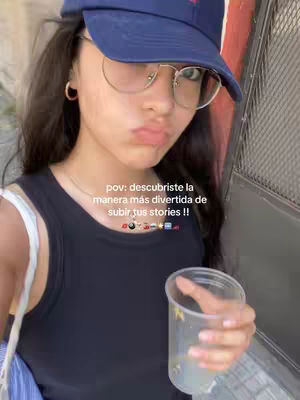 A young woman with dark hair and glasses looks towards the camera, pouting slightly. She wears a blue baseball cap and a black tank top. She holds a clear plastic cup in her right hand. The background appears to be outdoors, possibly a partial view of a building.