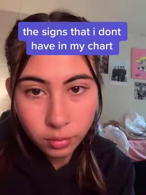 A selfie-style shot with a woman in the center. A text overlay in a blue box above her head reads, \"the signs that I dont have in my chart.\" She has brown hair, and dark eyes. The background is simple, appearing to be a room interior possibly a bedroom, with items partially visible, suggesting a casual setting. The lighting is soft.