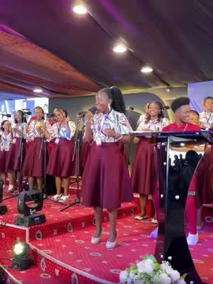 The cover image shows a well-lit stage with red carpet and a backdrop with several people standing on it. A woman in a burgundy skirt is at the fore, holding a microphone and singing. The stage is illuminated by overhead lights and a small floral arrangement at the base of the podium. The scene speaks to a performance, likely a church service or concert.