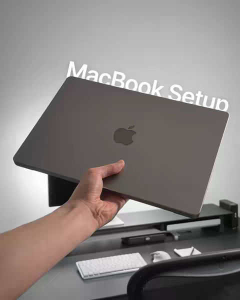 A person holding a gray laptop with the Apple logo against a gray background, revealing a desk setup. White text overlays the laptop, and the overall composition suggests a focus on tech or productivity.