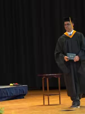 A man in a graduation gown and cap walks on a wooden stage, holding a document. The background is a dark curtain.