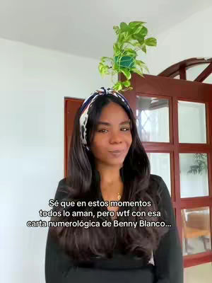 A woman stands in front of a white wall with a door behind her. She has long dark hair, a headband, wearing a black top and a small necklace. A hanging plant sits atop her head. Text appears on the screen.