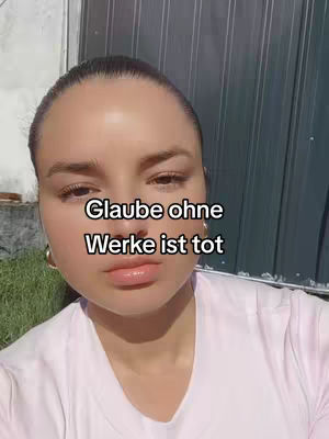 A close-up selfie of a woman against an outdoor background. She has light skin, dark hair pulled back, and is wearing a pale pink shirt and gold hoop earrings. The background is a mix of a green grassy area and a dark panelled wall. Text overlayed on the image reads 'Glaube ohne Werke ist tot'.