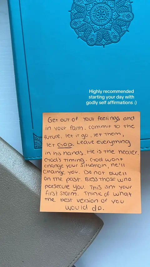 The image showcases a light blue leather-bound notebook with a decorative circular pattern embossed on the cover. A small, orange sticky note is placed on top, filled with handwritten black text. Beside this, a section of a gray notebook can be seen. The overall composition is simple, with a focus on the handwritten note.