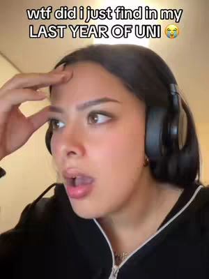 A young woman with dark hair wearing headphones looks surprised, touching her forehead with one hand and with her mouth open as if speaking or gasping. The background is lightly blurred and appears to be indoors.