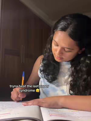 A woman with curly dark hair is shown in a dimly lit room, bent over a notebook, writing with a pen. The image is framed from a medium angle, focusing on her upper body and the desk space she is using. The lighting is soft, possibly from a nearby lamp, casting a gentle glow on her face and the notebook. In the bottom left corner of the image, a white text overlay reads "tryna beat the last paper syndrome" in a casual sans-serif font. The room's background includes a dark wooden cabinet.