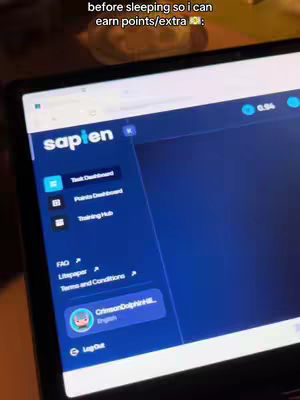 A person is using a laptop in a dark setting. The laptop screen displays a website interface with the logo "sapien" at the top left corner. Interface elements include a task dashboard, points dashboard, and training hub. The screen is angled, suggesting the person is holding the laptop. The environment is dimly lit, with a yellow glow from an undetermined source.