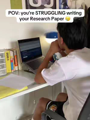 A young man is hunched over a laptop at a desk, with a laptop and open book in front of him, along with some other books and some stationary to his left. He is wearing a white t-shirt and black shorts. The text overlay reads 'POV: you're STRUGGLING writing your Research Paper'.