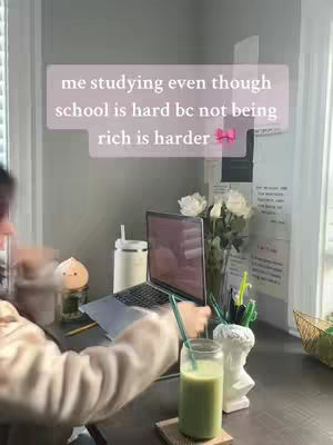 A well-lit workspace is shown, with a laptop centrally placed. The laptop screen is visible while a person's arm is reaching towards a glass of green drink. A glass is on a wooden coaster. Other items include a lamp, vase with white flowers, and notes on the wall.