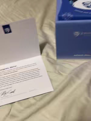 The image shows an interior shot with a bed covered in a neutral-toned fabric. A white acceptance letter on a white background is open towards the left of the frame; the text inside the letter is too small to read. A blue box with the John Hopkins University logo on it lies in the background, also out of focus; one of the crops hints that the box might have contained dorm information within it.