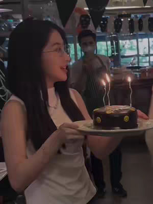 The cover image shows a woman in a white top holding a plate with a small chocolate cake decorated with three lit candles. She is smiling and looking to the side. A person wearing a white shirt and apron is in the background, along with a partially visible interior setting. The overall lighting is neutral, with a focus on the woman and the cake. The scene appears to be taken indoors.