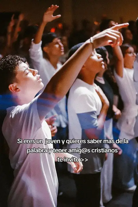 The image shows a group of young adults gathered in what seems to be a church or religious setting. The focus is on a young man in the foreground, seen from the chest up, who is raising his right arm and pointing upward with his right index finger. His eyes are closed, and his face shows an expression of earnestness or devotion. A group of people are visible in the background, with some raising their hands in a similar gesture. The lighting appears to come from above, highlighting the central figure. Overlay text at the bottom of the image, in a white, simple font, invites the viewer to join if they're interested in learning more about the word and having Christian friends.