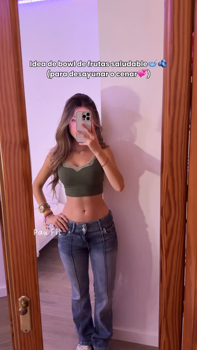 A woman is standing in front of a mirror, holding a phone up to take a selfie. She is wearing a green crop top, blue jeans, and bracelets. The background is a bedroom with a white wall and a wooden door frame.