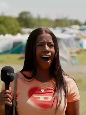 A woman with long black hair stands in an outdoor setting, holding a microphone. She is wearing an orange Diesel t-shirt with a red heart design. Her mouth is wide open as if she is speaking or surprised. Behind her, a blurred background suggests a park or outdoor event with tents visible.