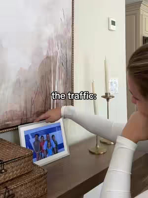 A woman is leaning towards a digital photo frame, pointing to the family photo displayed on its screen. The frame sits on a wooden surface below a large abstract painting, above a decorative basket. The woman is wearing a long-sleeved white shirt. The overlay text appears in the top - The Traffic: