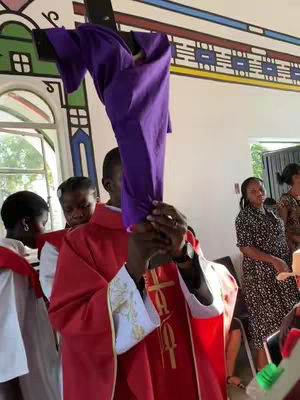 A priest dressed in a red cassock holds a purple cloth-draped cross high in a church setting. Several people, including young girls, are in the background, suggesting a religious ceremony. The image has a documentary feel, with natural lighting and focus.
