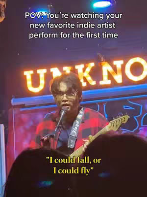 The cover image shows a live performance scene in a dimly lit setting. A person wearing glasses and a plaid shirt is performing, playing a guitar and singing into a microphone. Text overlays at the top and bottom frame the scene. The lighting is warm, and the background features a lit-up sign.