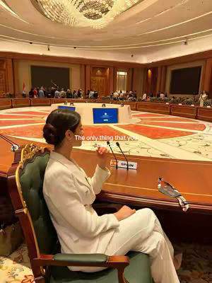 A woman in a white suit sits pensively at a large, round conference table. The setting appears to be a formal meeting room, with a large chandelier and other people in the background. The woman is the central focus, with the text overlay conveying a motivational message.