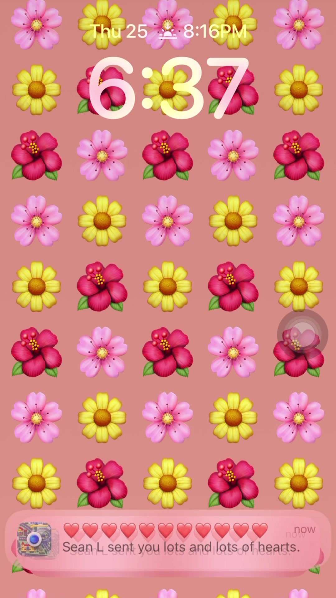 A phone screen displaying a lock screen with a pattern of stylized flowers in various colors. The top of the screen shows the time and date, and a notification bubble at the bottom displays a message and multiple heart emojis.