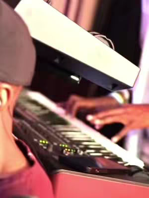 The image shows a close-up of a person playing a keyboard. The focus is on the musician's hands and the keyboard itself, with blurry background elements. The lighting is soft and warm.