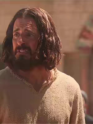 The image shows a close-up of a man with long, dark hair. He has a beard and a slightly pained expression, suggesting sadness. He appears to be in an outdoor setting, with warm, brown tones in the background.