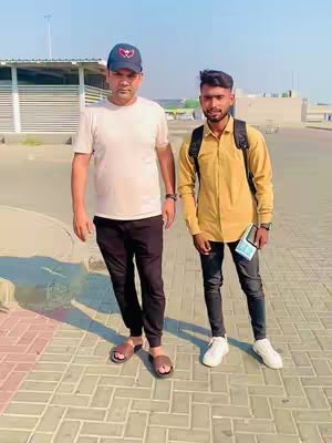 Two men standing on a paved area near a building and a fence, likely at an airport. The person on the left is dressed in a white t-shirt, black pants, and sandals, while the person on the right has a yellow shirt, black jeans and white sneakers.