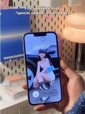 A hand holding a smartphone displaying an incoming call from someone named Jennie. The phone screen shows a picture of Jennie in a car setting. The background includes a textured wall, shelves with items like a blue album, and a lamp.