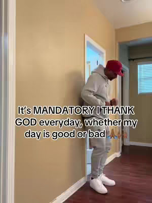 A man in a grey hoodie and sweatpants, wearing a red baseball cap, stands indoors leaning against a beige wall, near an open doorway. Text overlay reads: "It's MANDATORY I THANK GOD everyday, whether my day is good or bad".