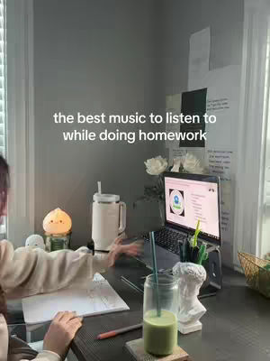 A person with dark hair is sitting at a desk, facing left, with a laptop open. The laptop screen displays content. A glass of green drink and a pen sit on the desk. The background includes a window and some white paper with writing on the wall.