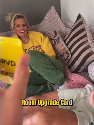 A woman with blonde hair smiling on a bed with pillows in the background. A yellow card with “RVSD” on it is held in the foreground towards the camera and text overlay “Room Upgrade Card”.