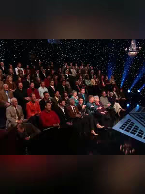 The image captures a large gathering seated in an auditorium-like setting, likely for a performance or event. The audience is densely packed, dressed in a mix of formal and business casual attire. The lighting is dramatic, with spotlights illuminating the stage and the audience in a darkened room. The background features a starry design, and a section of stage is visible on the right. The overall composition suggests a live event, possibly a concert or a religious gathering.