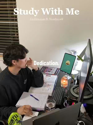A young man is seated at a desk, engrossed in studying. The foreground includes a laptop, open books with a pen resting on a notepad, and a glass of water. A digital tablet on a stand displays visual icons. The background is a plain wall, a window with blinds and a computer.
