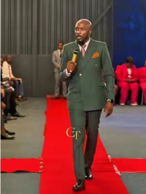 A man in a green suit stands on a red carpet, holding a microphone. He appears to be addressing an audience. The background includes other individuals, seated and standing.