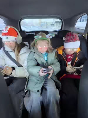 Three children sitting in the back seat of a car. They are all wearing winter coats and Santa hats or reindeer antlers. Each child is looking at their phone, and there is a filter over their eyes. The background is blurred, showing the outside of the car.