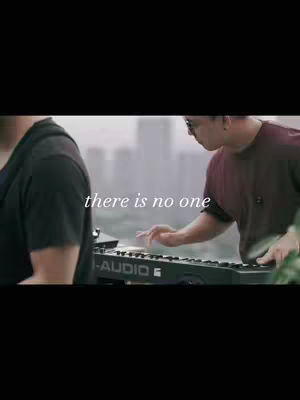 The image shows a musical performance. Two men are visible, with one on the left partially cut off, dressed in a dark shirt. The man on the right, wearing a maroon shirt and glasses, is playing a keyboard, with a cityscape blurred in the background. The text overlay "there is no one" is placed on top.