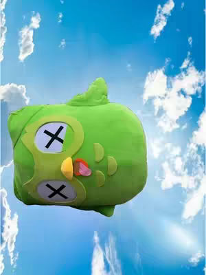 A green, plush owl toy with white eyes crossed out with black 'X's. It's set against a vibrant blue sky with scattered white clouds. The owl has a yellow beak, a pink tongue and some green details on the side. The overall composition is playful and eye-catching.
