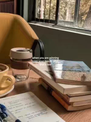 An indoor shot of a desk setup with a book titled \"Journal\" open on the wooden desk. A stack of books, cup of coffee or tea, and a clear glass with a beige lid and a straw inside are also visible. The background includes a window and a chair.