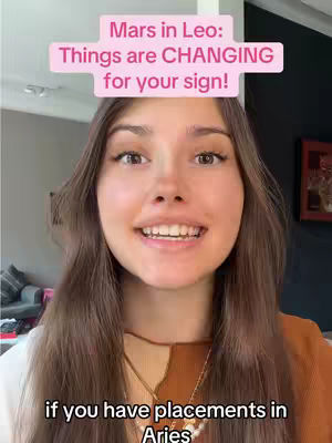 A young woman with long brown hair is centered in the frame, smiling warmly. A pink banner reading 'Mars in Leo: Things are CHANGING for your sign!' sits above her head. Below, text states, 'if you have placements in Aries.' The background is a blurred interior.