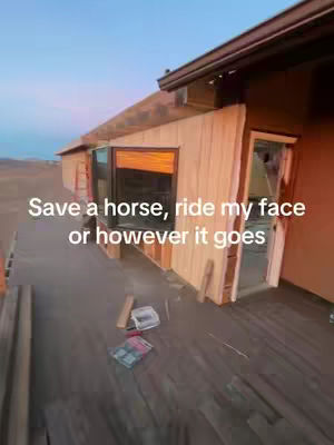 An angled exterior shot of a house under construction. The main focus is on a section of the building with unfinished wooden siding and an open doorway. The background shows a dry, possibly desert landscape under a blue sky.