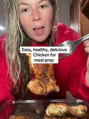 A woman with long blonde hair is holding a piece of cooked chicken with a fork, presenting it to the camera. The chicken is seasoned with a dark spice blend and is held over a clear glass dish also containing chicken. The background is slightly blurred, showing a kitchen setting with wooden cabinets.