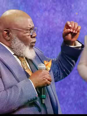 A man in a patterned blazer and gold tie with a microphone in his hand, talking.