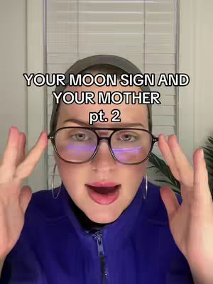 A woman with glasses holds them up with her hands, looking at the camera with an open mouth. Above her is white text that reads "YOUR MOON SIGN AND YOUR MOTHER pt. 2". The background includes a window with blinds.